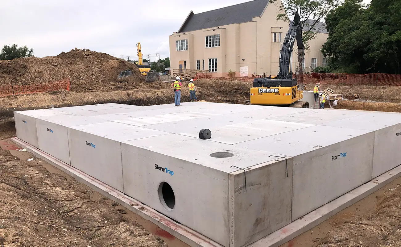 Construction workers stand on top of a large, rectangular concrete StormTrap stormwater detention system for churches in an excavated pit next to a yellow excavator.