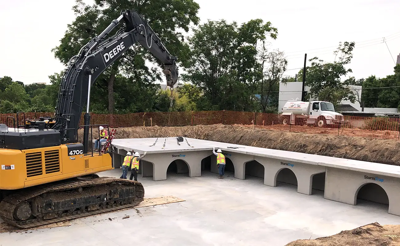 A yellow excavator lowers a large concrete StormTrap culvert section for a stormwater detention system for churches into an excavation pit where workers are positioned.